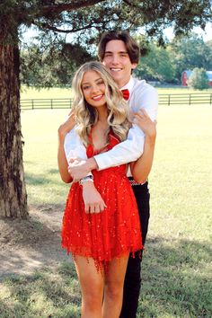 a man and woman standing next to each other in front of a tree with their arms around each other