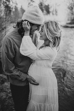 a pregnant couple standing next to each other in front of a river and trees, with the man kissing the woman's forehead