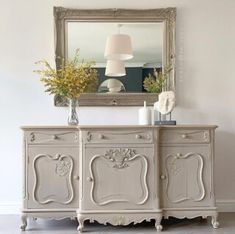 a white dresser with flowers on top in front of a mirror and lamp above it