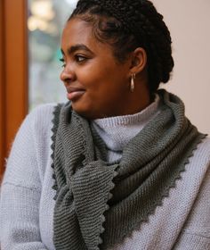 a woman with braids wearing a gray sweater looks off to the side while sitting in front of a window