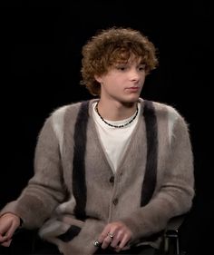 a young man with curly hair sitting in a chair and wearing a striped cardigan