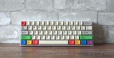 a computer keyboard sitting on top of a wooden table next to a brick wall with colorful keys