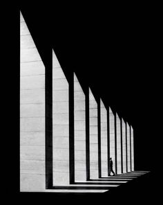 black and white photograph of person walking in front of wall with long shadows on it