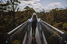 a woman walking across a bridge in the middle of some trees with clouds above her