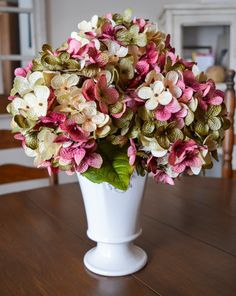 a white vase filled with lots of pink and green flowers on top of a wooden table