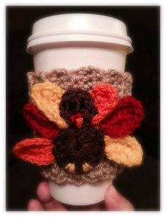 a coffee cup with a crocheted turkey on it's side and a hand holding the cup