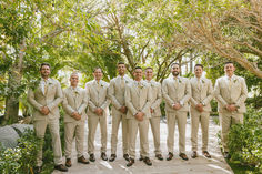 a group of men standing next to each other in front of trees and bushes at a wedding