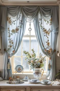 a kitchen window with flowers on the curtains and dishes in front of it, next to a potted plant