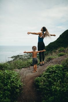 a woman and child standing on top of a dirt road near the ocean with their arms outstretched