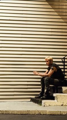 a man sitting on top of a bench next to a tall metal building with a wall in the background