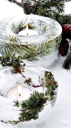 two candles are sitting in the snow next to pine cones and evergreen needles, with one candle lit