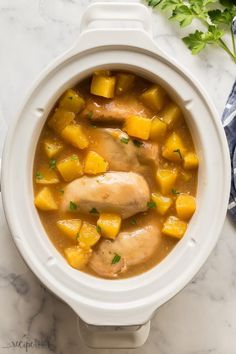 a white crock pot filled with chicken, potatoes and parsley on top of a marble counter