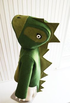 a stuffed animal that is wearing a green dinosaur costume with eyes and tail, standing in front of a white wall