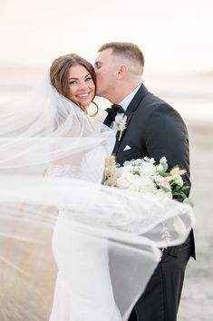 a bride and groom kissing on the beach with their veil blowing in the wind behind them