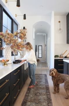 a woman standing in a kitchen next to a dog on a rug and looking at the floor