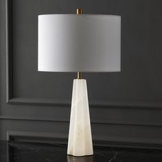 a table lamp with a white shade sitting on top of a black table next to a gray wall