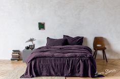 a bed with purple sheets and pillows in a white room next to a wooden chair