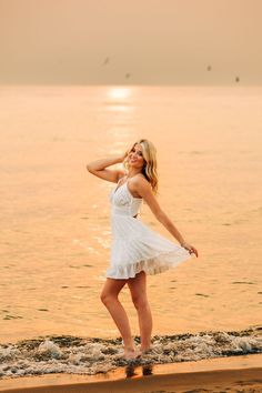 a woman in a white dress standing on the beach with her hands behind her head