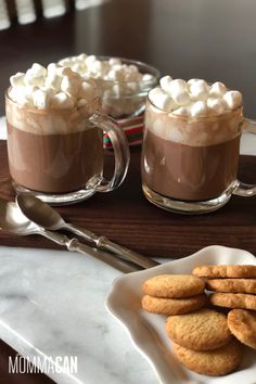 hot chocolate with marshmallows in two mugs on a plate next to cookies