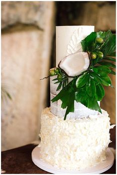 a three tiered white cake with green leaves and coconuts on the top layer