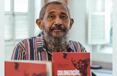 an old man sitting at a table with two books in front of him and the book cover is red