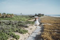 a bride and groom walking down a path by the beach with an umbrella over their heads