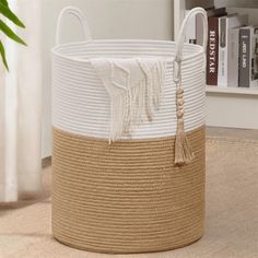 a white and tan basket with tassels on the bottom is sitting in front of a bookshelf