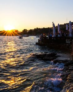 people are sitting on the dock watching the sun go down over the water as boats pass by