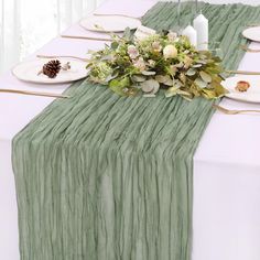 the table is set with white plates and green napkins on it, along with greenery