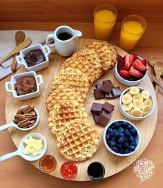 a wooden plate topped with waffles, fruit and other foods next to glasses of orange juice