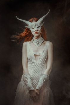 a woman with red hair wearing a white mask and dress, standing in front of a dark background