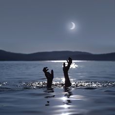 two people reaching up into the air in water at night with moon and mountains in background