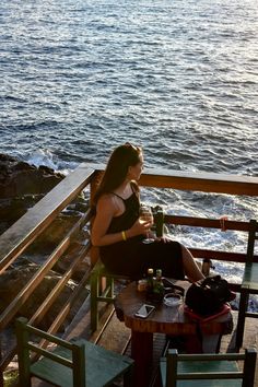 a woman sitting at a table on top of a wooden deck next to the ocean