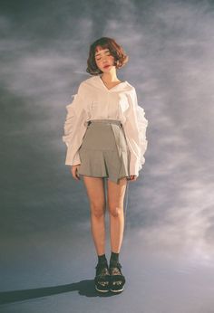 a woman standing in front of a gray background wearing shorts and a white shirt with ruffle sleeves