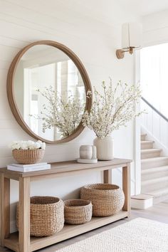 a wooden table with baskets and flowers on it in front of a round mirror above the stairs