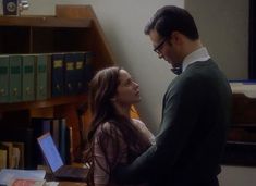 a man and woman standing next to each other in front of a desk with books on it