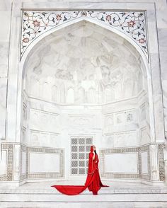 a woman in a long red dress is standing near a white wall with an arch