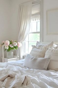 a white bed topped with lots of pillows and blankets next to a vase filled with flowers