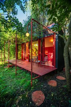 a small red cabin in the woods surrounded by green grass and trees, with a wooden deck