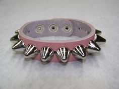 "1 Row Large 1/2\" UK/77 Apex Cone Studs Genuine Leather Handcrafted Bracelet This a handcrafted, genuine leather 3/4\" wide Cuff/bracelet. The Leather is nice and soft, it will get buttery and more subtle with wear. The leather is available in vibrant colors. Racing Red Midnight Black Pretty Pink Arctic White Please check select color at time of check out. The studs/spikes are large UK77 Cone studs. The pyramids studs measure 1/2\" in diameter and conical shaped The Cone studs have a nice lustr Adjustable Edgy Pink Jewelry, Edgy Adjustable Pink Jewelry, Adjustable Leather Bracelet With Rivets For Concerts, Punk Jewelry With Rivets For Gift, Punk Jewelry With Rivets As Gift, Adjustable Rock Style Leather Bracelet For Gift, Adjustable Spiked Bracelets For Festivals, Punk Style Bracelets, Adjustable Punk Wristband With Spikes