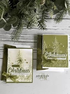 two christmas cards sitting on top of a table next to a pine tree and evergreen branches