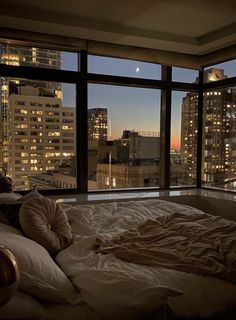 an unmade bed in front of a large window overlooking the city at night time