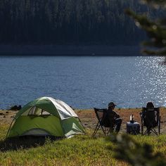 two people sitting in lawn chairs next to a tent on the shore of a lake