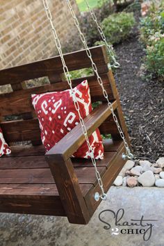 a wooden swing with two red pillows on it in front of a brick wall and stone walkway