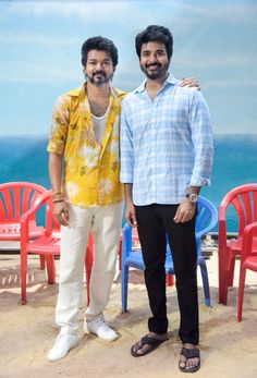 two men standing next to each other in front of red chairs and the ocean behind them