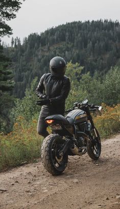 a man riding on the back of a motorcycle down a dirt road next to a forest