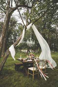 an outdoor table set up with white draping and flowers on it, next to a chandelier hanging from a tree