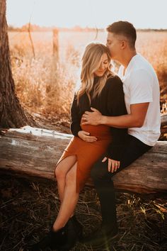 a man and woman sitting on a log in front of a tree with their arms around each other