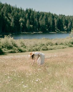 a woman in a white dress and straw hat crouches down to pick up flowers