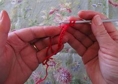 two hands are knitting red yarn on a bed sheet with floral print sheets behind them